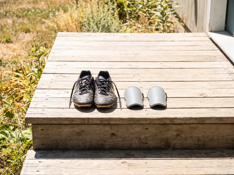 A pair of black youth soccer cleats and two shin guards side by side on a sunlit wooden porch step.